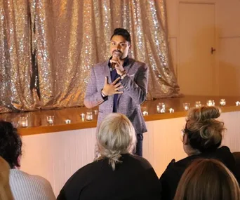Male speaker in a blazer performs on stage with candles, addressing a seated audience in a dimly lit room.