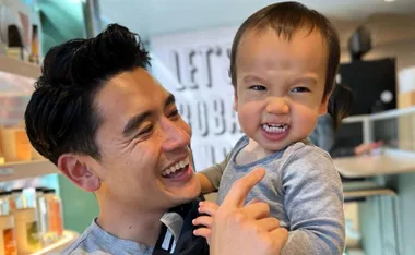 Father smiling at grinning toddler in a bright room, creating a joyful moment together.