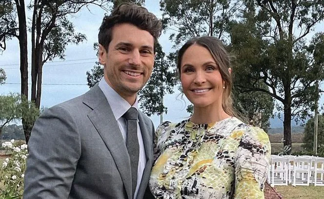 A couple smiling outdoors in formal attire, standing together with trees in the background.