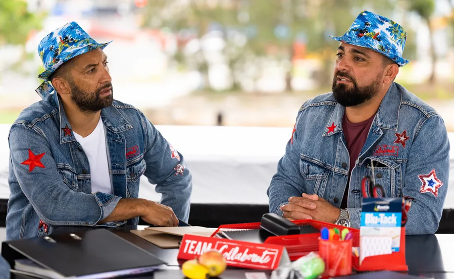 Two men in denim jackets and colorful hats are seated at a table with office supplies and a "Team Collaborate" sign.
