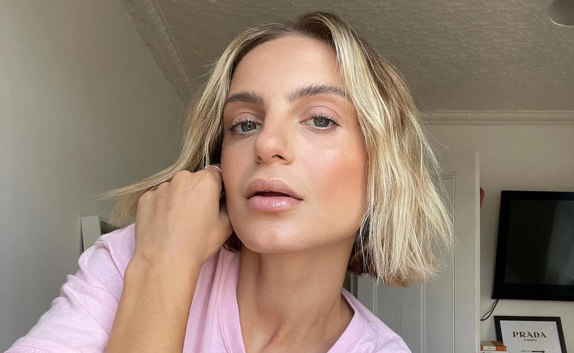 Blonde woman in a pink shirt taking a close-up selfie in a room with framed artwork on the wall.