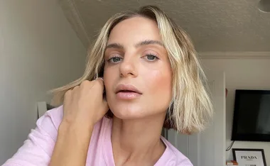 Blonde woman in a pink shirt taking a close-up selfie in a room with framed artwork on the wall.