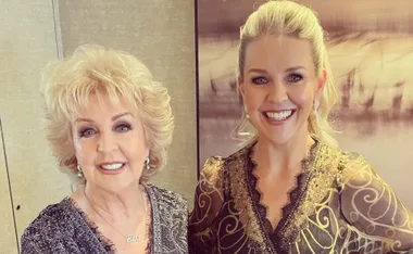 Two women smiling at the camera, dressed in intricate, elegant outfits.