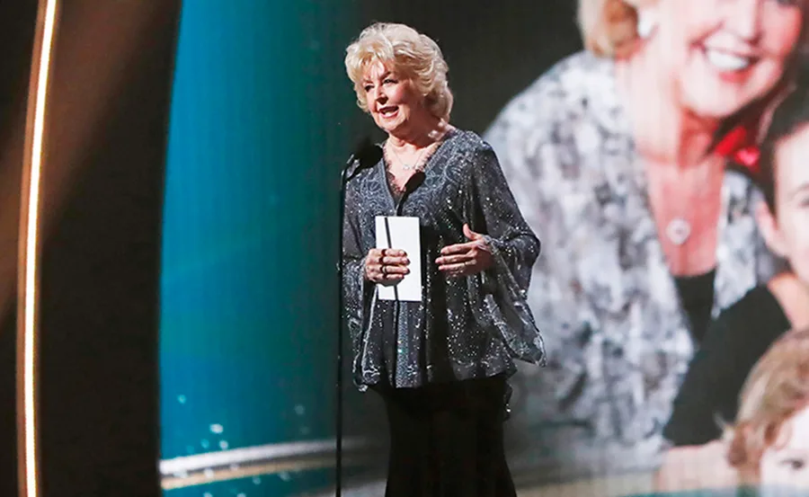 A person in sparkly attire speaks on stage at an awards event with a microphone and paper in hand.