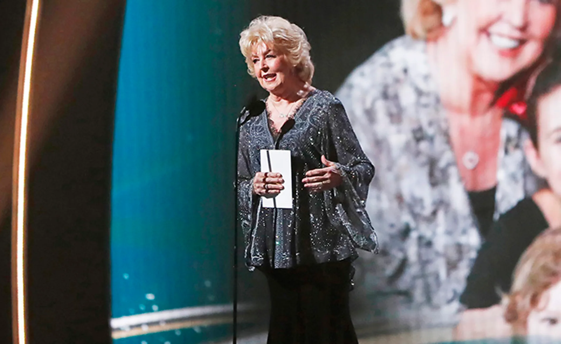 A person in sparkly attire speaks on stage at an awards event with a microphone and paper in hand.