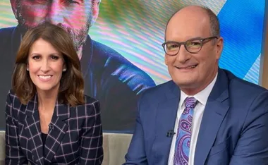 Two TV hosts smiling on set, with colorful background and a large screen behind them.