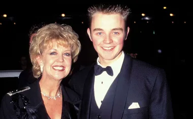 Two people smiling at an event; one in a tuxedo, the other in a black outfit with jewelry, night background.