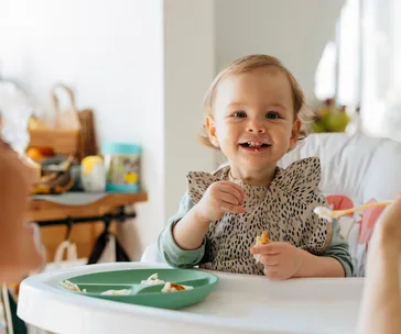 A toddler in a high chair eating, wearing a bib, with a plate of food on the tray, smiling at the camera.