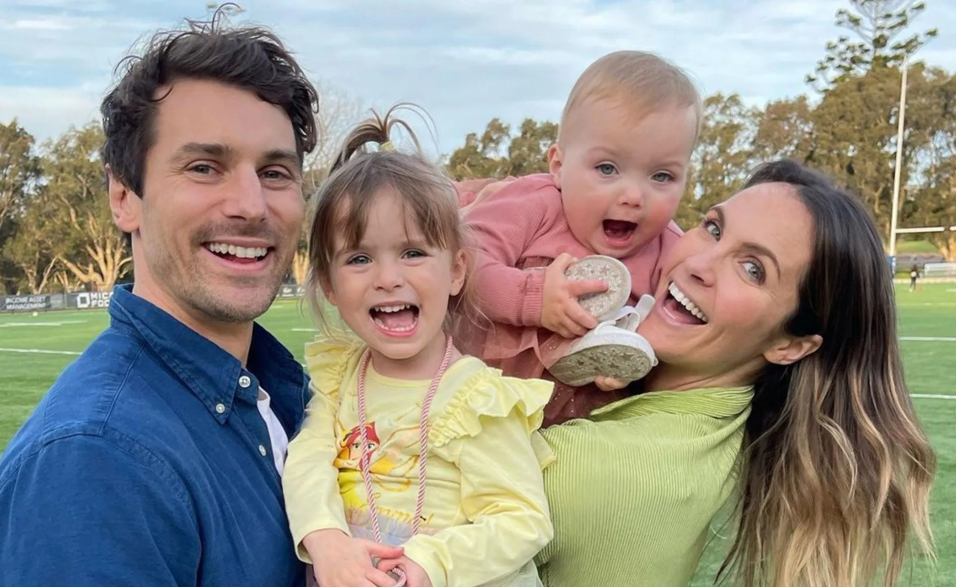 A family of four smiling outdoors; two parents holding two young children, with a grassy field in the background.