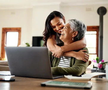 Granddaughter hugging grandmother as they smile at a laptop in a cozy home setting.