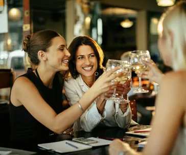 Women smiling and clinking wine glasses in a lively bar setting.