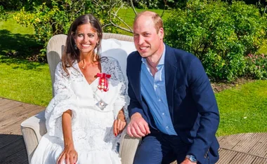 A woman in a white dress with honors sits next to a man in a blue blazer, both smiling in a green garden.