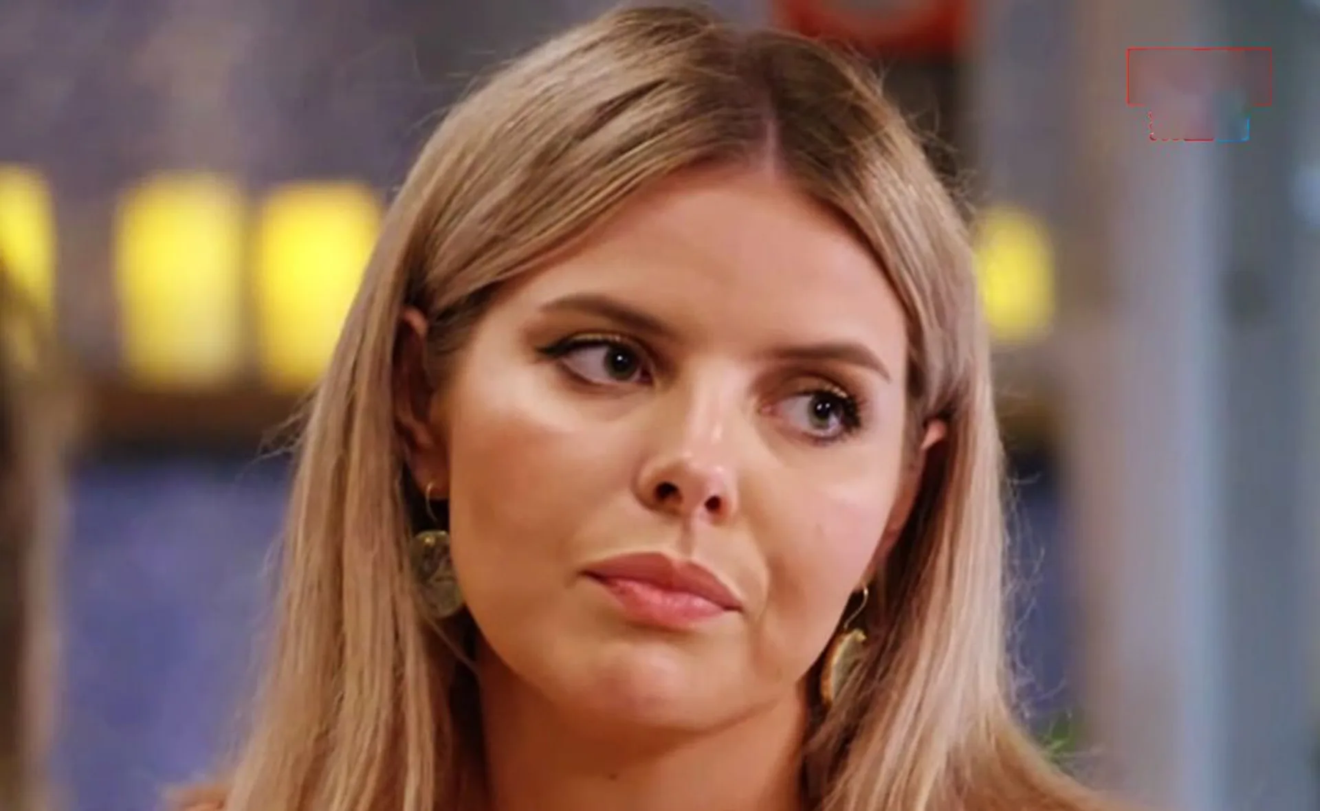 A woman with long blonde hair and earrings looks to the side, with blurred background and soft lighting.
