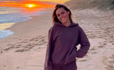 Person in a hoodie smiling on a beach at sunset, with waves and a vibrant sky in the background.