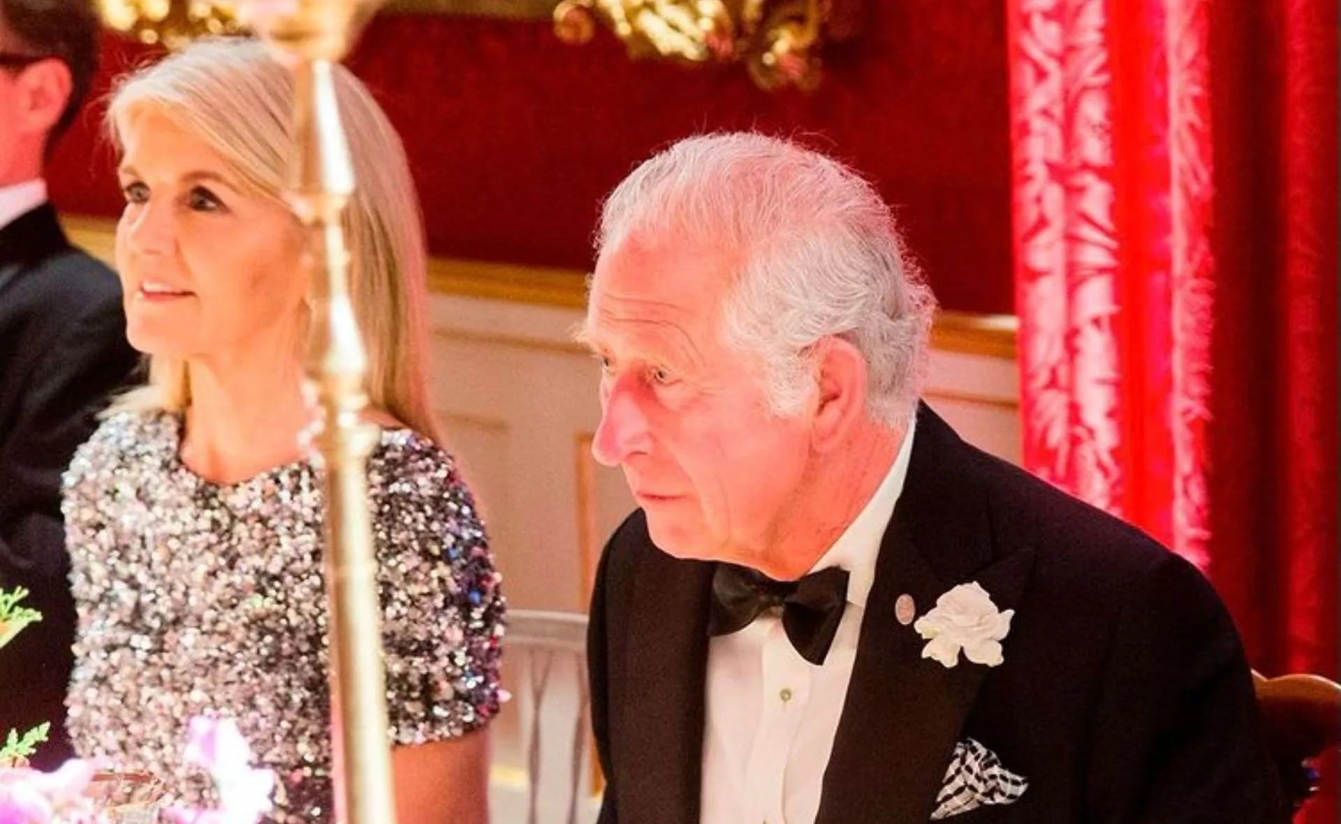 Two individuals seated at a formal event, man in tuxedo, woman in sparkly dress, red drapery in background.