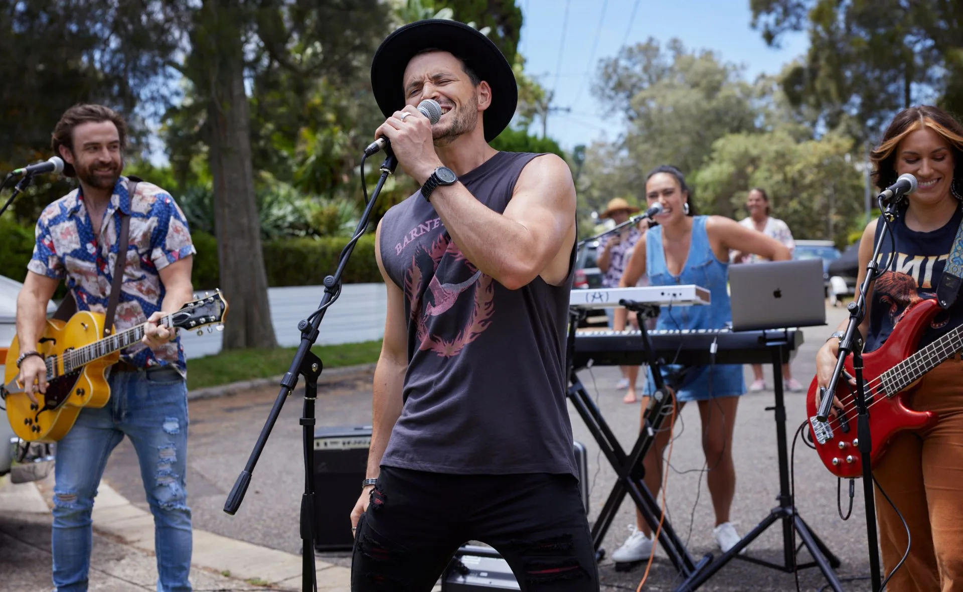 Band playing outdoors; lead singer in a sleeveless shirt and hat, with guitarist, keyboardist, and bassist in the background.