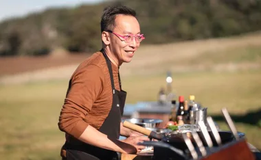 Chef wearing pink glasses and a brown shirt cooks outdoors, smiling, with utensils and ingredients on the counter.