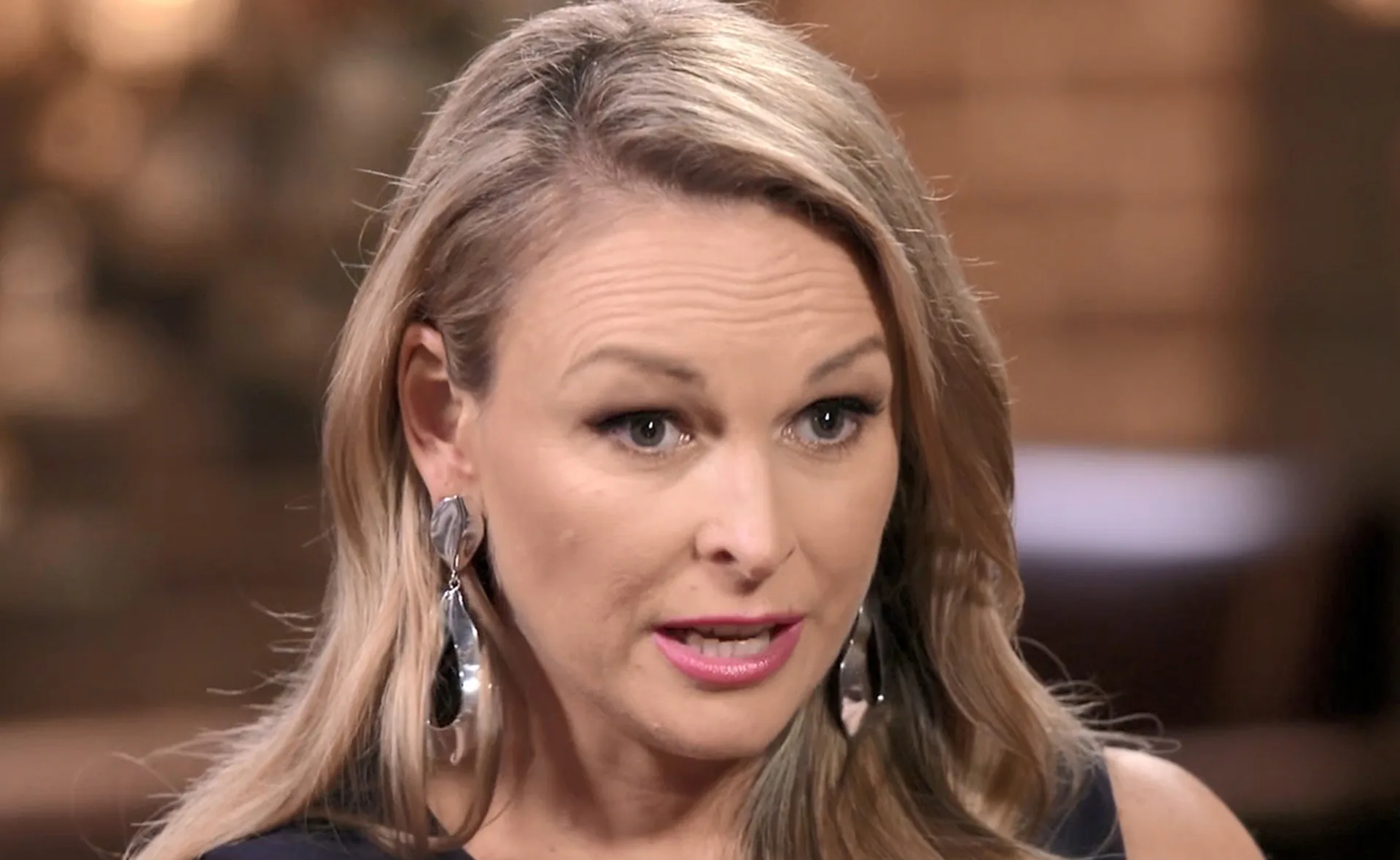 A woman with long blonde hair and dramatic earrings speaks intently in a dimly lit setting.