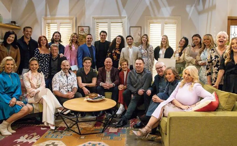 Cast of "Neighbours" finale gathered in a cozy living room, smiling and posing together.