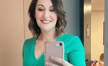 A person in a green outfit taking a mirror selfie with a phone, smiling warmly.