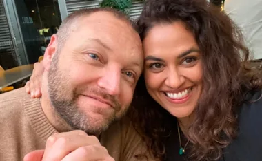 A couple smiling warmly at the camera, embracing each other closely in a relaxed setting.