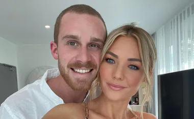 A couple smiling at the camera in a bright room with white curtains in the background.