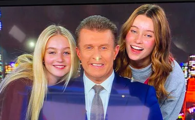 A man in a suit is smiling with two young women, both smiling, in a brightly lit setting with a cityscape backdrop.