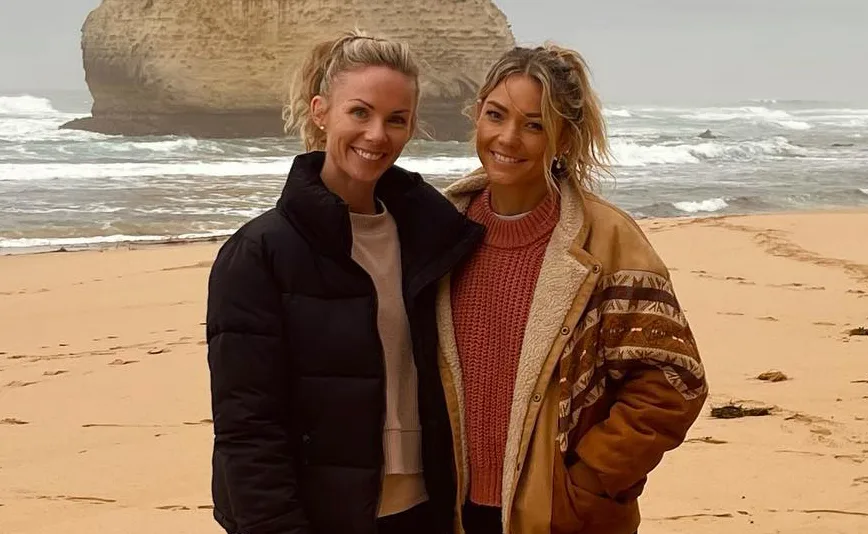 Two women smiling on a windy beach, wearing jackets, with waves crashing and a rock formation in the background.