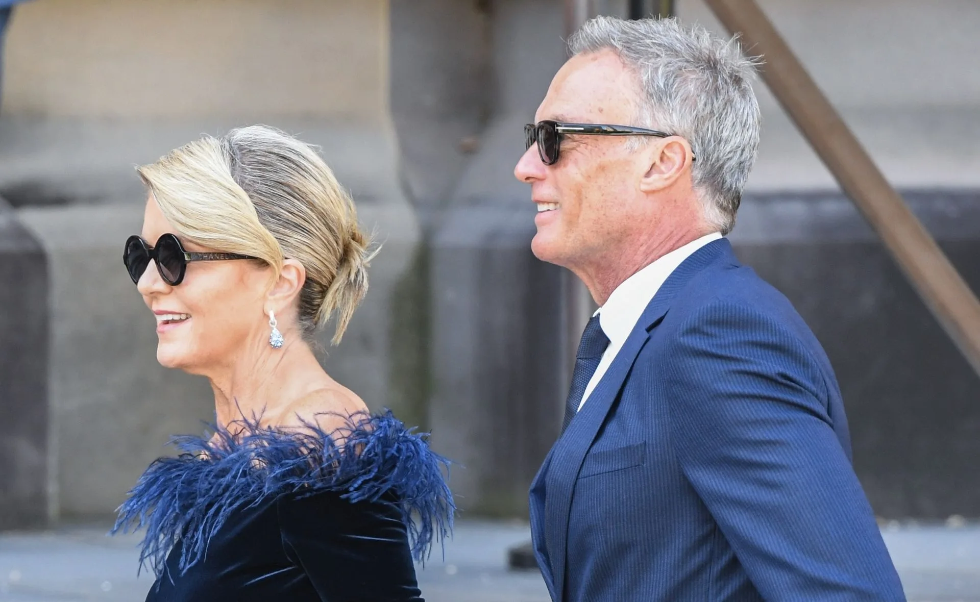 A man and woman in formal attire, both wearing sunglasses, walking side by side and smiling.