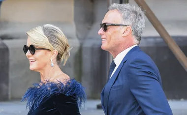 A man and woman in formal attire, both wearing sunglasses, walking side by side and smiling.