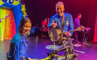 "Two performers in blue outfits on stage, one playing drums, the other with a banjo, smiling at the camera."