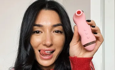 A person with long dark hair holding a pink Vush sex toy, playfully sticking out their tongue.