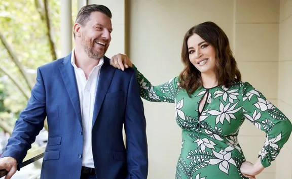 Two people on a balcony; man in blue suit laughing, woman in green floral dress smiling, trees in background.