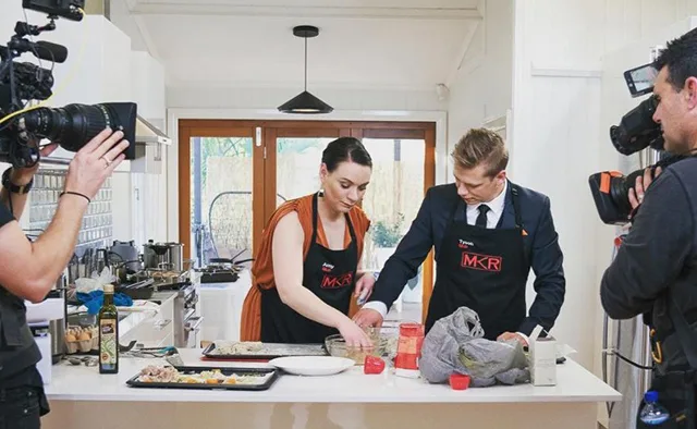Contestants on "My Kitchen Rules" prepare food in a kitchen, surrounded by camera crew filming the process.