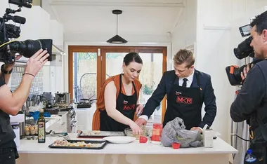 Contestants on "My Kitchen Rules" prepare food in a kitchen, surrounded by camera crew filming the process.