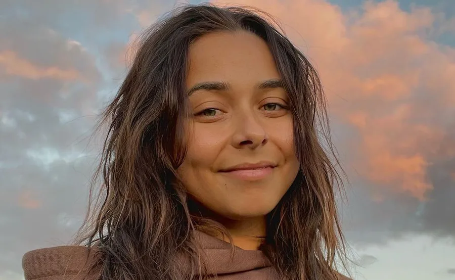 A person with long dark hair smiling under a cloudy sky during sunset.