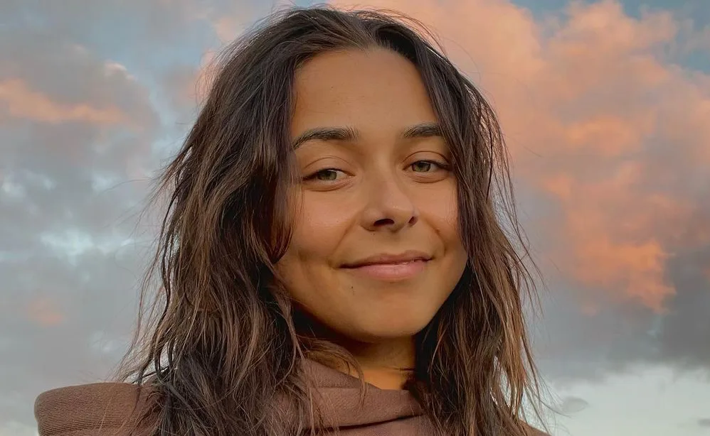 A person with long dark hair smiling under a cloudy sky during sunset.
