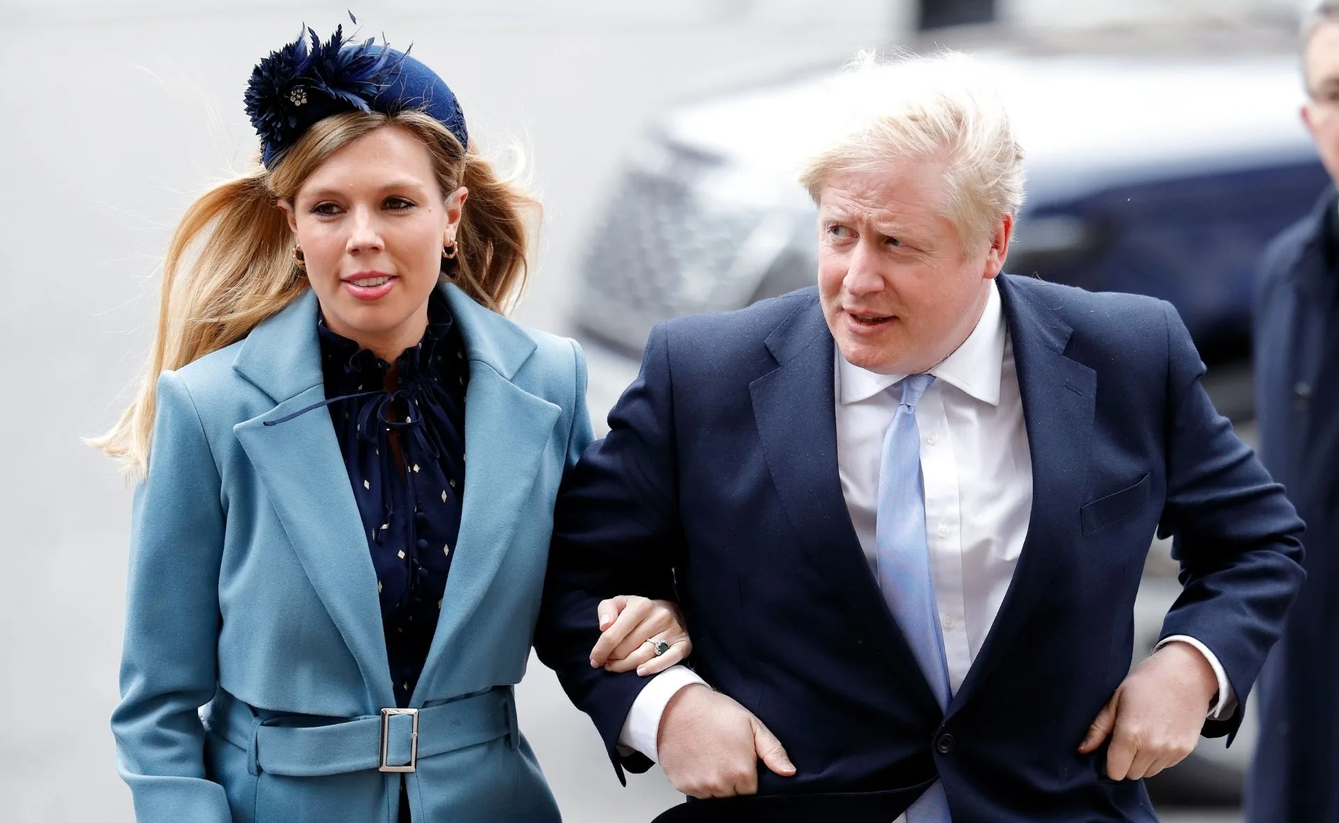 A man and woman walking together; he wears a suit, she wears a blue coat with a matching headpiece.