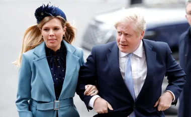 A man and woman walking together; he wears a suit, she wears a blue coat with a matching headpiece.