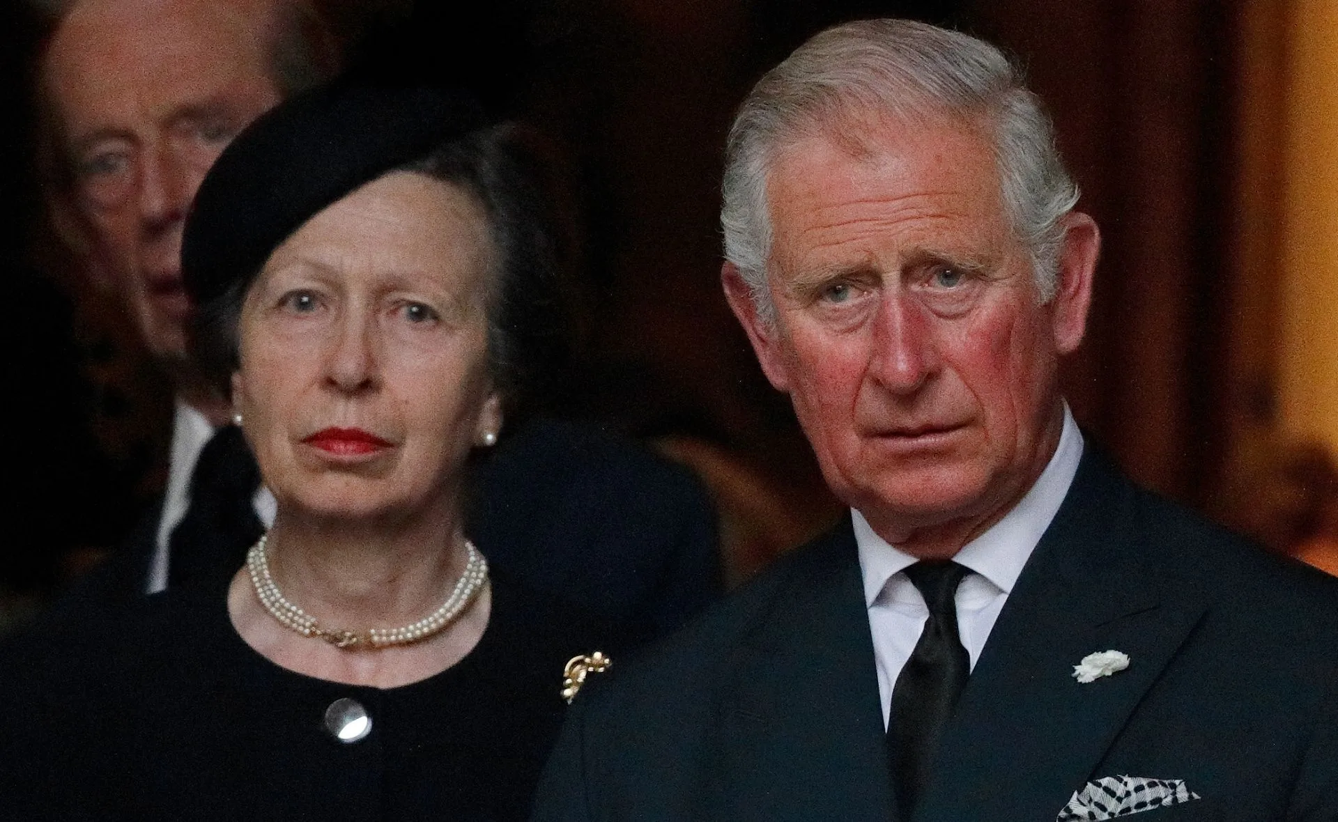 Two individuals formally dressed, standing closely, with serious expressions.