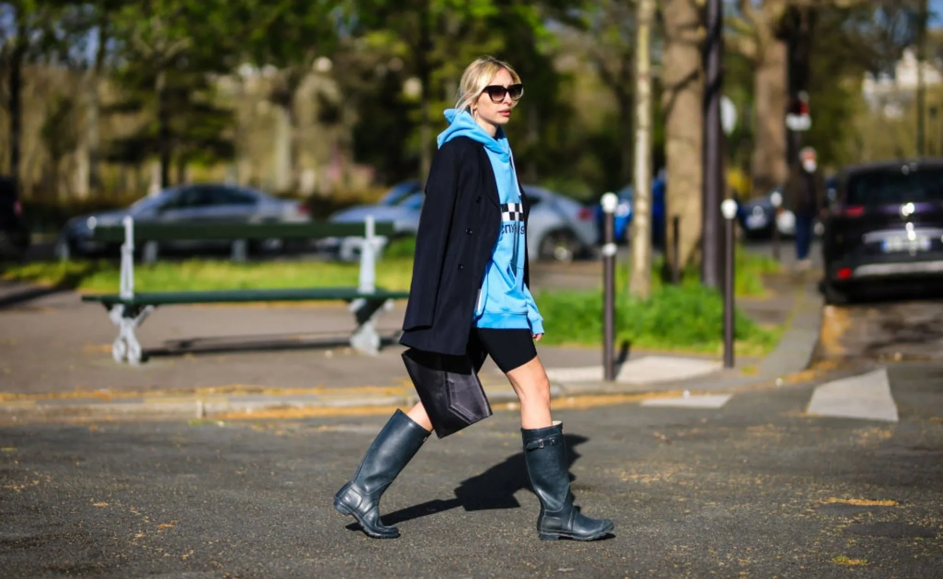 Woman in a blue hoodie, black coat, and waterproof boots walking in a park.