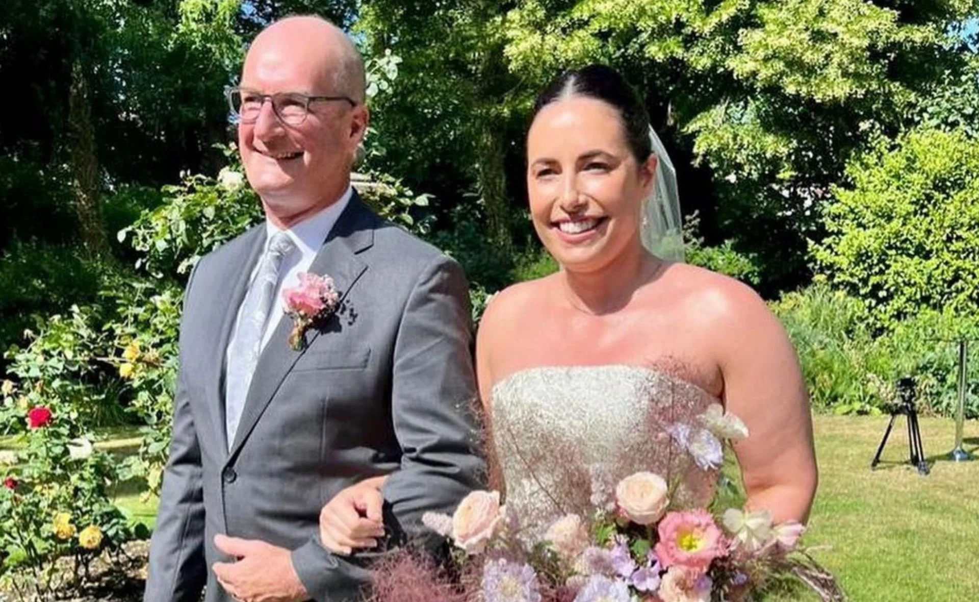 Father and bride walking arm-in-arm outdoors, surrounded by greenery and flowers.
