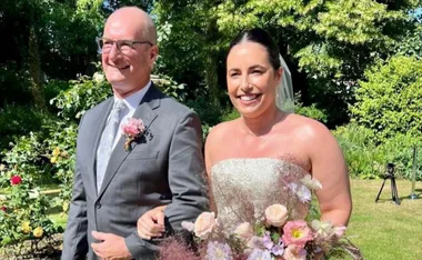 Father and bride walking arm-in-arm outdoors, surrounded by greenery and flowers.