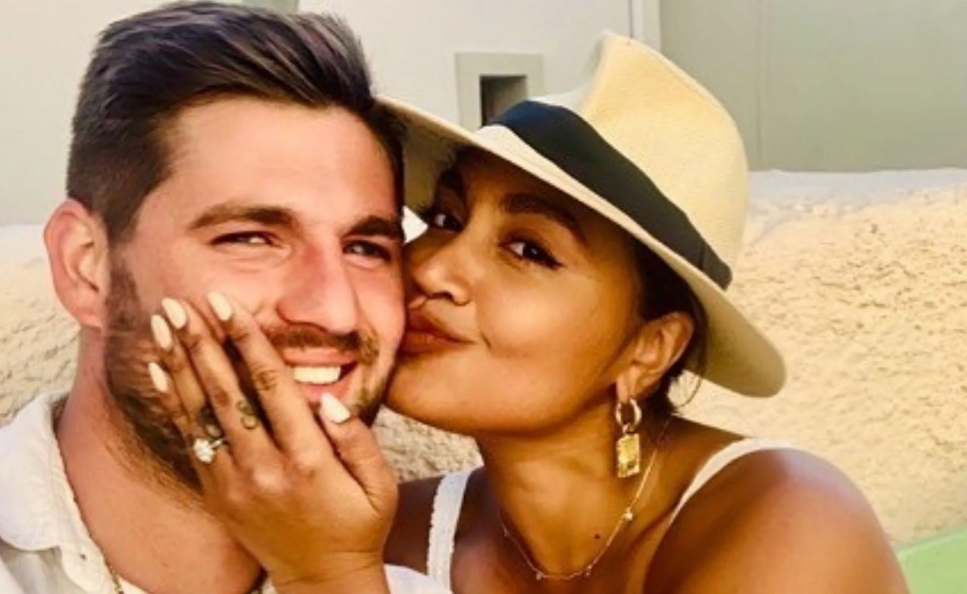 A woman in a hat affectionately kisses a smiling man, with her hand on his cheek displaying a ring.