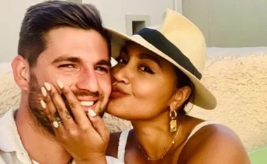 A woman in a hat affectionately kisses a smiling man, with her hand on his cheek displaying a ring.