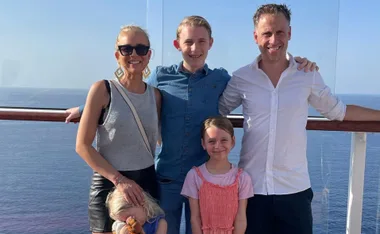 Family of five smiling on a deck, ocean in the background under a clear blue sky.