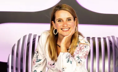 A woman in floral dress, smiling with her hand on her chin, sits against a modern, curved backdrop.