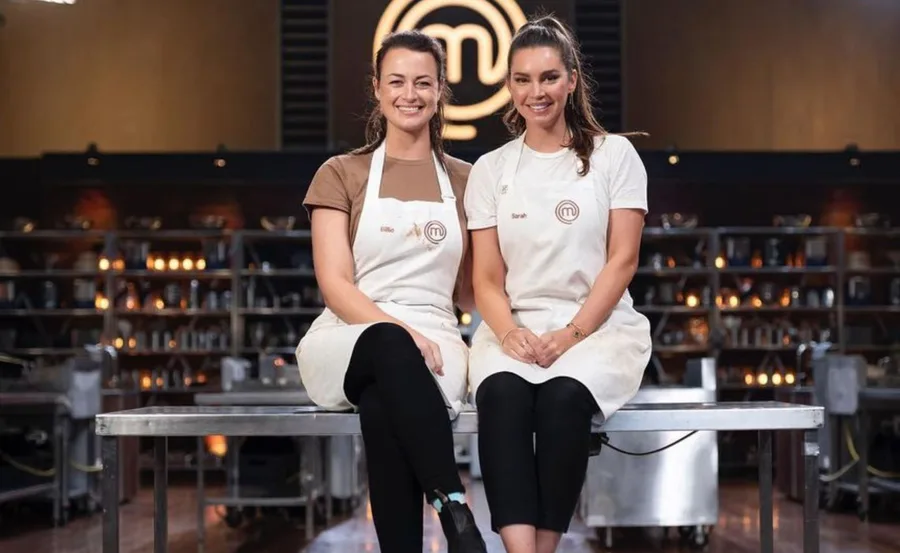 Two women in MasterChef aprons, Billie and Sarah, sitting in the MasterChef kitchen, smiling.