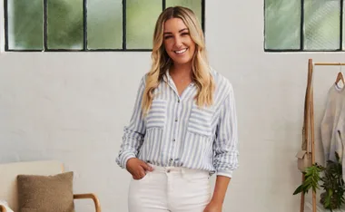 A woman with long blond hair is smiling, wearing a striped shirt and white pants, standing in a bright room.
