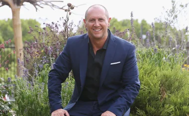Man in a blue suit smiling, sitting in a garden with colorful plants in the background.